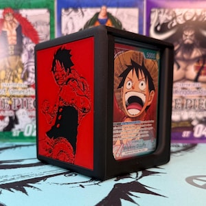 May include: Black card holder featuring a red Luffy design on one side and a card with Luffy's image on the other. The card holder is designed to protect and display trading cards. The background includes other trading card packs.