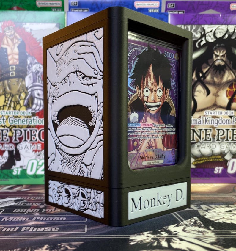 May include: A black and brown card deck box with a white and gray image of a character's face on the front. The box has a clear plastic window on the front to display a card. The card features a character with black hair and a red and blue outfit. The text on the card reads "Monkey D. Luffy". The text on the box reads "Monkey D."