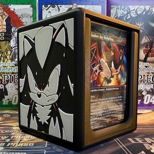 May include: A black and gold card storage box featuring a 3D embossed image of Sonic the Hedgehog. The box holds a card with the text "Shadow the Hedgehog". The box is designed to hold trading cards.