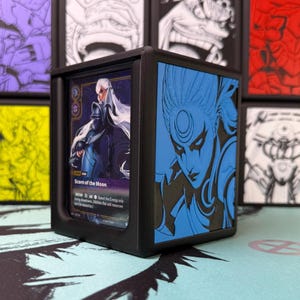 May include: A black 3D-printed deck box featuring a blue side panel with an embossed illustration of a character with a crescent moon symbol on their forehead. A trading card titled Scorn of the Moon is visible inside the front-facing slot.