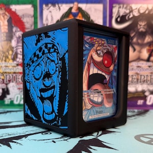 May include: A black display case holding a trading card featuring a cartoon clown with a large red nose and open mouth. The case has a blue graphic on the side and is surrounded by other trading cards.