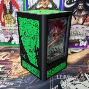 May include: Black card storage box with green accents and a green illustration of a character. The box has a clear window displaying a card with the text "Roronoa Zoro". The box is designed for trading card games.