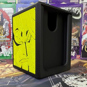 May include: Black card storage box with a yellow embossed design on one side. The design features a character's face. The box has a rectangular shape with a curved opening on one side. The box is designed for trading card games.