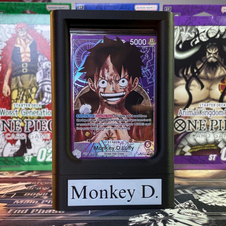 May include: A black card protector case with a clear plastic window displaying a collectible trading card featuring Monkey D. Luffy from the One Piece anime series. The card has a purple and blue background with the text "5000" and "Monkey D. Luffy" printed on it. The case has a black label on the bottom with the text "Monkey D."