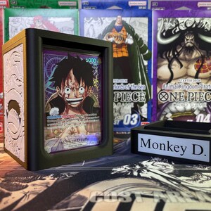 May include: A white and gold deck box with a black border and a clear plastic window showing a One Piece trading card featuring Monkey D. Luffy. The box is sitting on a table with other One Piece trading cards in the background. The text on the card reads "Monkey D. Luffy".