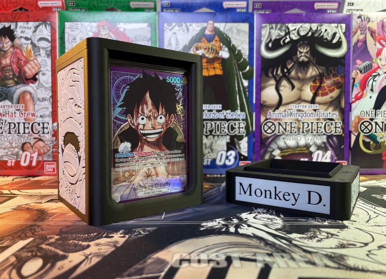 May include: A white and gold deck box with a black border and a clear plastic window showing a One Piece trading card featuring Monkey D. Luffy. The box is sitting on a table with other One Piece trading cards in the background. The text on the card reads "Monkey D. Luffy".