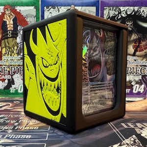 May include: A black card display case with a yellow embossed demon design on the side. The case holds a trading card with a monster illustration. The card has the number 5000 printed on it. The case is designed to protect and showcase collectible cards.