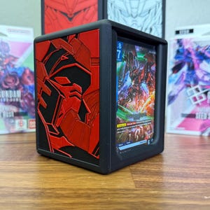 May include: Black card holder with a red and black embossed design of a robot's face. The holder displays a colorful card with a Gundam character. Other Gundam-themed products are visible in the background.