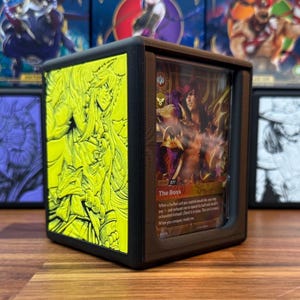 May include: A black display case with a card inside, featuring a character illustration. The case has a yellow side with a detailed relief design. The card reads "The Boss" with text below. The case is designed to protect and showcase collectible cards.