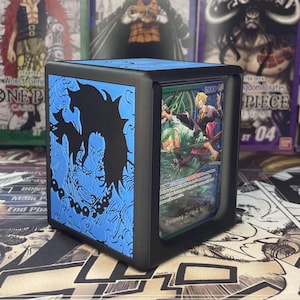 May include: Black and blue card storage box featuring a character design on the side. The box holds a trading card with a green and yellow illustration. The box has a black frame and a blue top.