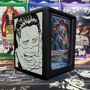 Puede incluir: Caja de almacenamiento de cartas negra con una ilustración detallada en blanco y negro de la cara de un personaje en la parte delantera. La caja contiene una carta coleccionable con una ilustración colorida. La caja está diseñada para el juego de cartas One Piece.