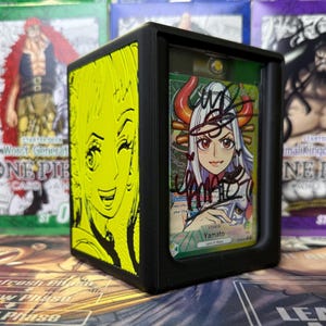 May include: Black card holder with a yellow embossed anime character on the side. It displays a signed Yamato card from the One Piece card game. The card is visible through a clear window.