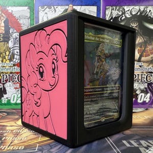 Puede incluir: Caja de almacenamiento de cartas negra y rosa con una imagen en relieve rosa de Pinkie Pie. La caja contiene cartas coleccionables, una de las cuales es visible, con un personaje guerrero. La caja está diseñada para proteger y exhibir cartas coleccionables.