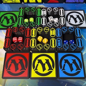 May include: A collection of black plastic game token holders with colored accents in green, red, yellow, and blue. Each holder features circular slots for dice and text labels like Vigilance, Lifelink, Trample, and Hexproof. Includes matching square coasters with a stylized M logo.