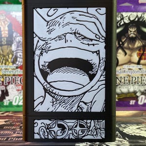 May include: Black card game deck box with a white and black illustration of a character's face with a large open mouth. The bottom of the box has a white and black illustration of a pair of glasses.