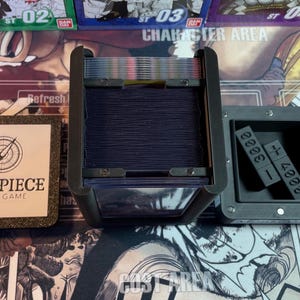 May include: A black card game box with a clear plastic lid, showing a deck of cards inside. The box has a label that says "One Piece Card Game". There is a separate black box with a compartment for dice.