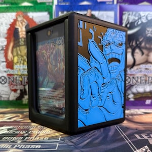 May include: Black card storage box with a blue embossed image of a character's face and hand. The box holds a card with the number 5000 visible. The box is designed to protect trading cards.