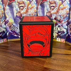 Op de afbeelding: Een zwarte deckbox met een rode en zwarte illustratie van een personage uit de anime-serie One Piece. De doos heeft de tekst "One Piece Card Game" aan de zijkant.