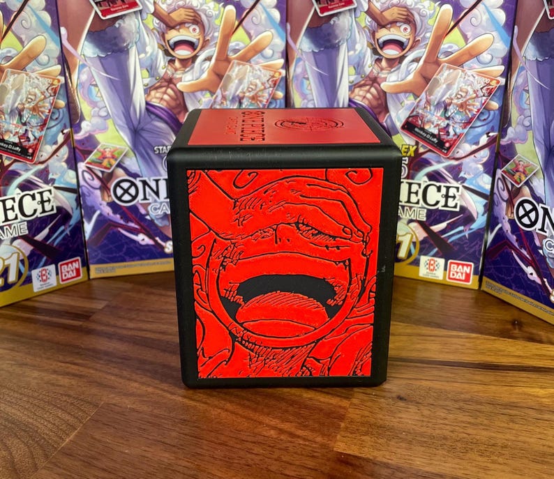 Op de afbeelding: Een zwarte deckbox met een rode en zwarte illustratie van een personage uit de anime-serie One Piece. De doos heeft de tekst "One Piece Card Game" aan de zijkant.