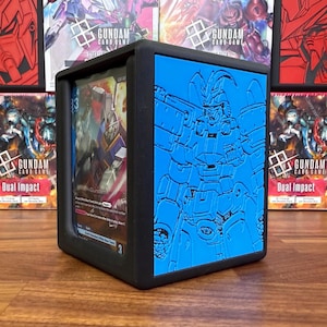 May include: Black card storage box featuring a blue embossed Gundam design. The box displays Gundam trading cards through a clear panel. Gundam card game boxes are visible in the background.