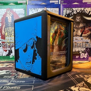 May include: A black and gold card storage box with a blue embossed image of a character's face on the side. The box is open, revealing a card with a character in a green coat. The background shows several unopened card game decks.