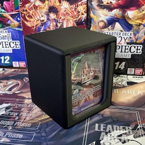 May include: A black deck box with a clear window showing a trading card featuring a character with red hair and a green bandana. The card has the text "Monkey D. Luffy" and "5000" on it.