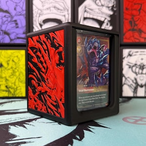 Puede incluir: Una caja de almacenamiento de tarjetas de plástico negro con un panel lateral rojo texturizado que presenta un diseño en relieve intrincado y de bordes afilados. La abertura frontal muestra una carta coleccionable.