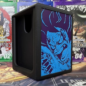 May include: Black card storage box featuring a blue monster design on a purple panel. The box has a slot for easy access and is surrounded by other card game boxes.