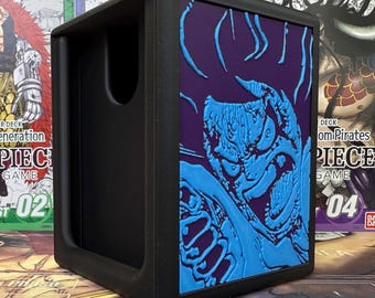 May include: Black card storage box with a blue monster design on a purple background. The box has a slot for easy access and is surrounded by other card game boxes.