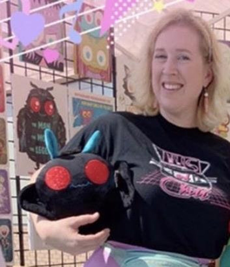 K&ouml;nnte beinhalten: Eine Person tr&auml;gt ein schwarzes T-Shirt mit einem pinkfarbenen und wei&szlig;en Aufdruck eines futuristischen Fahrzeugs und dem Wort "TRUST". Die Person h&auml;lt ein schwarzes Pl&uuml;schtier mit roten Augen und blauen Antennen.