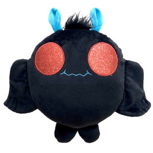 K&ouml;nnte beinhalten: Ein schwarzes Pl&uuml;schtier in Form einer Fledermaus mit roten Glitzeraugen und blauen Antennen. Das Spielzeug hat ein kleines, gesticktes L&auml;cheln.