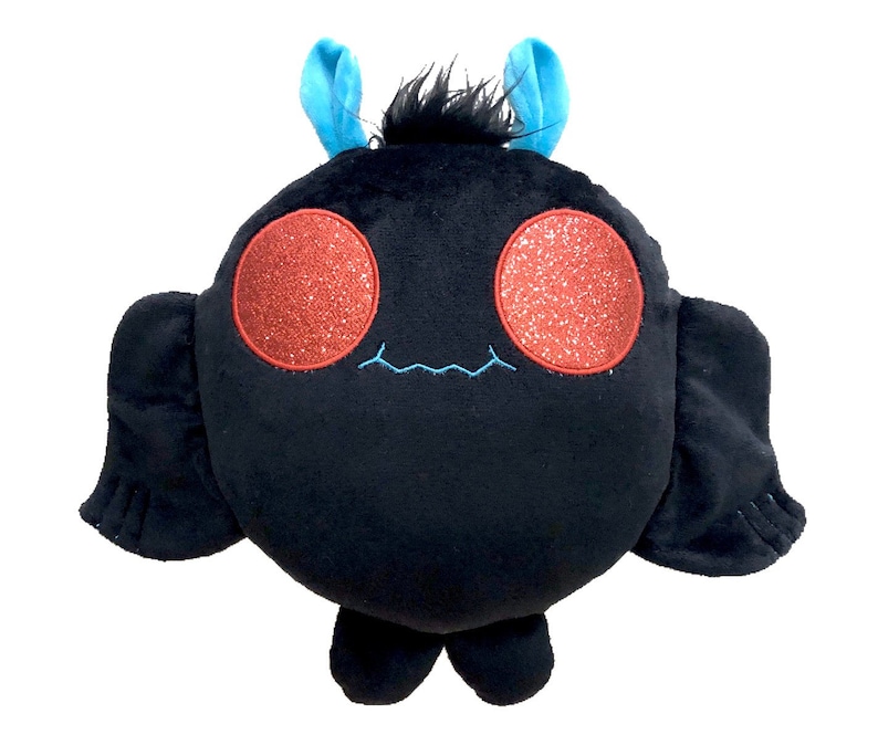 K&ouml;nnte beinhalten: Ein schwarzes Pl&uuml;schtier in Form einer Fledermaus mit roten Glitzeraugen und blauen Antennen. Das Spielzeug hat ein kleines, gesticktes L&auml;cheln.