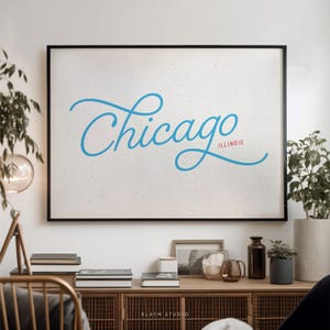 May include: A framed print with the word "Chicago" in light blue script, with "Illinois" in red below it. The print is set against a textured white background and framed in black. The print is displayed above a wooden cabinet.