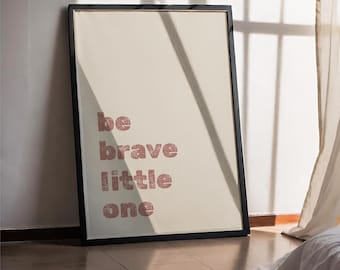 Puede incluir: Una impresión enmarcada con las palabras "be brave little one" en una fuente marrón rojiza texturizada. La impresión está sobre un fondo color crema y enmarcada en negro. La impresión está apoyada contra una pared.