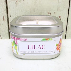 May include: A silver tin candle with a clear lid. The candle is white and has a wooden wick. The label on the tin is white with pink, yellow, and orange flowers. The label reads "Lilac 100% Soy Candle Hand Poured Vegan."