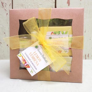 May include: A gift box with a yellow ribbon and a tag that reads "Lemon Verbena." The box is brown and contains yellow products, including soap and a jar. The products are from Green Daffodil and handmade in Michigan.