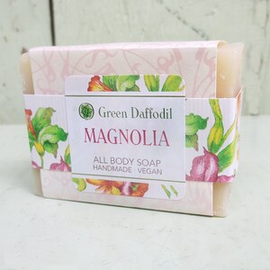 May include: A bar of Magnolia all-body soap wrapped in a decorative paper band. The band features floral illustrations in green, pink, and yellow, with the words "Green Daffodil" and "Magnolia" printed on the front. The soap is handmade and vegan.