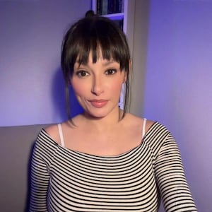 Może przedstawiać: Osoba z ciemnymi włosami i grzywką, ubrana w czarno-biały top w paski z odkrytymi ramionami. Osoba patrzy prosto w kamerę. Tło to jednolita fioletowa ściana.