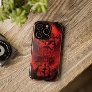 May include: A red and black phone case featuring a detailed graphic design. The case has a dark red background with a silhouette of a figure and architectural elements. The design is accented with black and green details. The phone case is on a wooden surface.