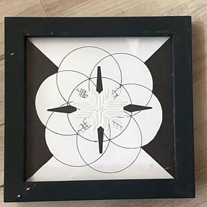Può includere: Un'opera d'arte astratta incorniciata con una cornice nera. L'opera presenta un design geometrico in bianco e nero con cerchi sovrapposti e un motivo centrale a forma di stella. Il design include forme nere a forma di freccia.