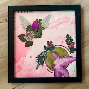 Può includere: Opera d'arte quadrata incorniciata con uno sfondo acquerello rosa. L'opera presenta elementi floreali e simili a insetti in viola, verde e bianco. La cornice è nera.