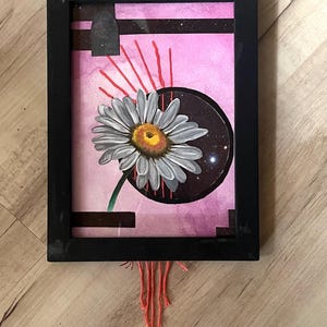 Può includere: Opera d'arte incorniciata con una grande margherita con petali bianchi e centro giallo. Lo sfondo rosa presenta un cerchio scuro con stelle. Linee rosse irradiano dal cerchio e fili arancioni pendono dalla cornice nera.