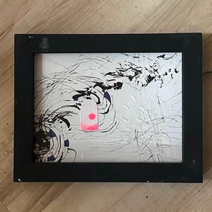 Può includere: Opera d'arte astratta in una cornice nera. L'opera presenta uno sfondo bianco con pennellate nere e blu e una forma rettangolare rosa. La cornice è nera e rettangolare.