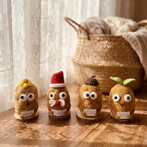 Op de afbeelding: Vier handgemaakte gehaakte aardappel figuren met wiebelogen en kleine houten voetstukken. Eén draagt een kerstmuts, een ander een bloem, een derde een bruine pet en de laatste groene bladeren. Elk heeft een klein label met tekst.