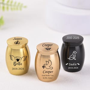Puede incluir: Tres pequeñas urnas de metal dorado y negro con nombres y fechas grabados. Las urnas son para cenizas de mascotas y presentan una paloma, un perro y un ángel con un corazón.