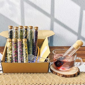 May include: A tea gift set with glass tubes containing dried herbs and flowers, sealed with corks. A cup of tea steeps next to the box, with a tea infuser and a wooden coaster. The box is brown and the tea is a deep red colour.