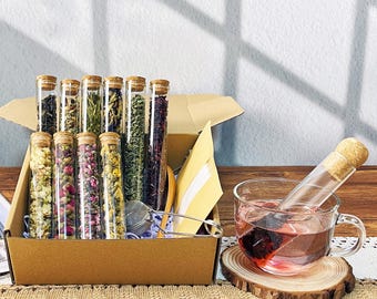 May include: A tea gift set featuring glass tubes filled with various dried herbs and flowers, sealed with corks. A cup of tea steeps next to the box, with a tea infuser and a wooden coaster. The box is brown and the tea is a deep red color.