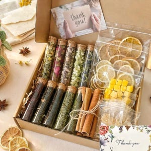 Pode incluir: Uma caixa de presente cheia de chá e itens relacionados. Contém tubos de vidro com ervas secas, rodelas de frutas cítricas secas e paus de canela. Inclui um cartão com a frase "Thank you!"