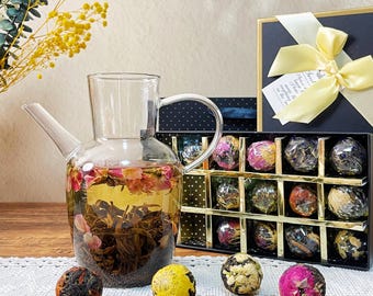 May include: A clear glass teapot filled with tea and flower petals, next to a box of tea balls. The tea balls are in various colors, and the box is tied with a yellow ribbon. A sprig of yellow flowers is in the background.