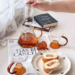 Puede incluir: Una tetera de vidrio y dos tazas de té con asas y bases de color ámbar, llenas de té. Un pequeño frasco de vidrio con hojas de té, un libro con la frase "I have a dream" y un plato con dos sándwiches.